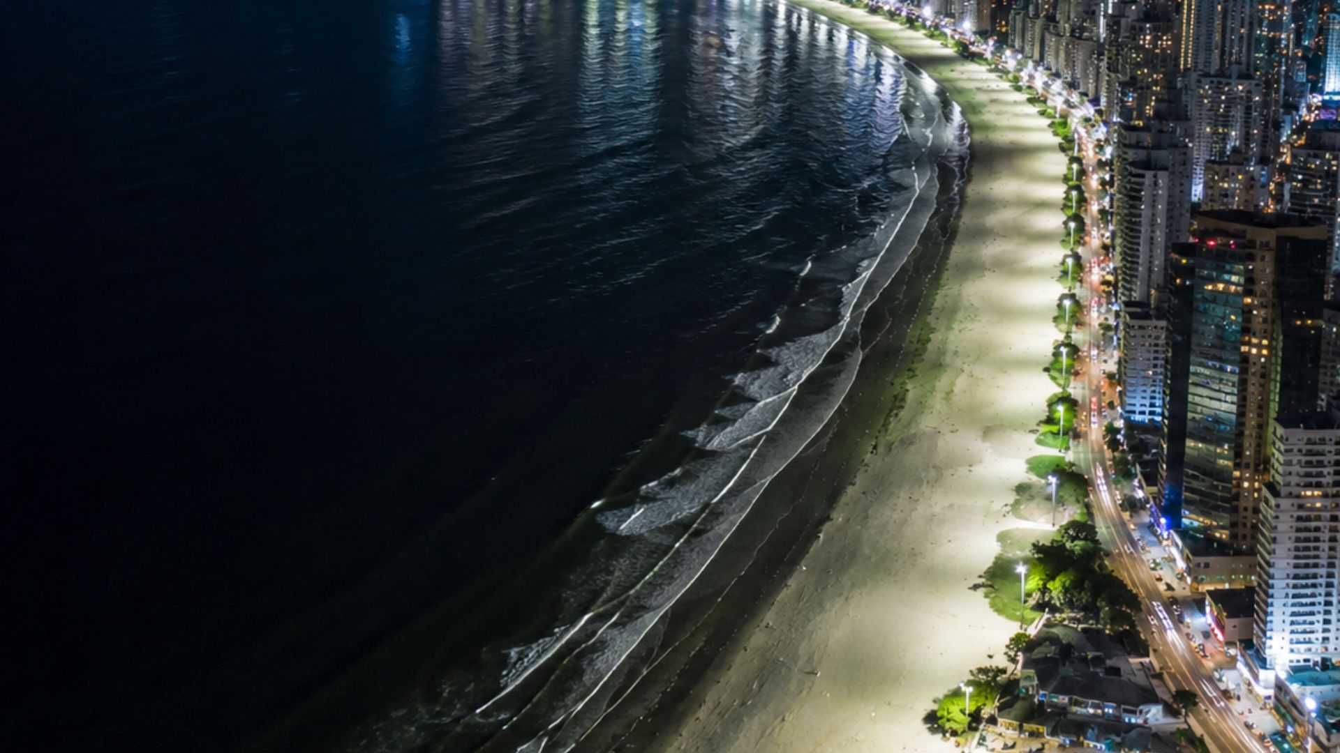 Vista aérea noturna de orla urbana litorânea com calçadão iluminado, avenida movimentada e prédios altos residenciais e comerciais à direita, mar escuro refletindo as luzes da cidade à esquerda, paleta azul-escura e dourada, iluminação noturna dramática, ângulo alto, fotografia realista de drone