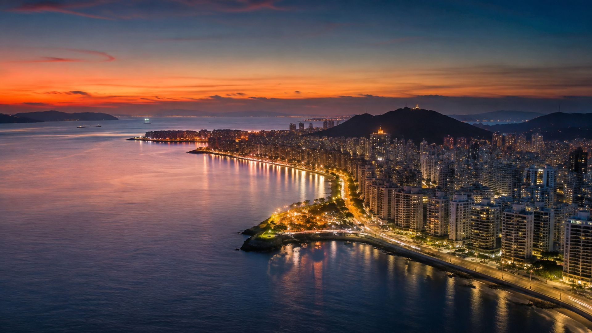 Vista aérea noturna da orla de Vitória, Espírito Santo, com prédios iluminados à beira-mar, céu em degradê laranja e azul no entardecer, reflexos dourados na água, fotografia realista panorâmica